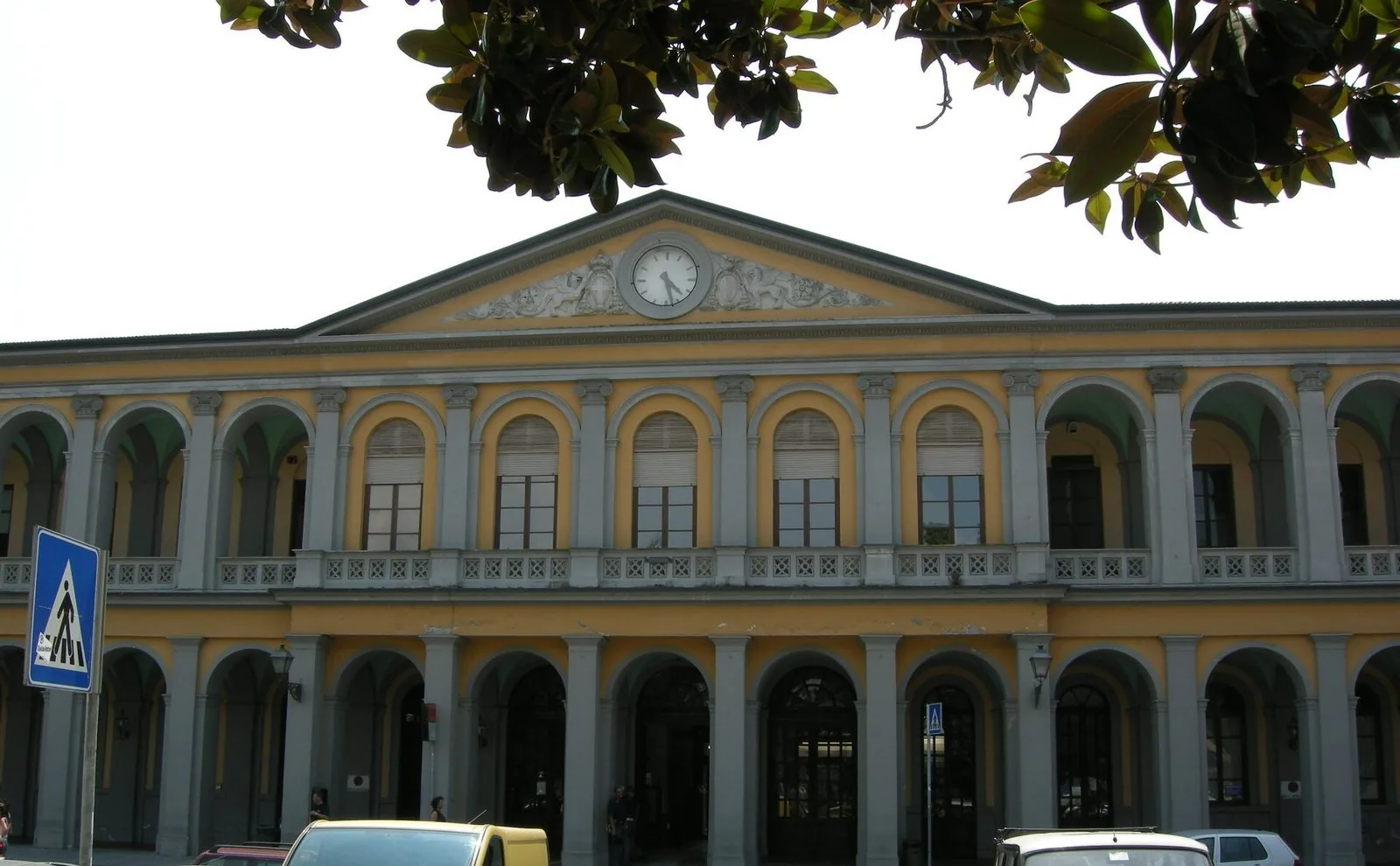Facciata porticata gialla e grigia della stazione ferroviaria di Lucca con l'orologio sul timpano